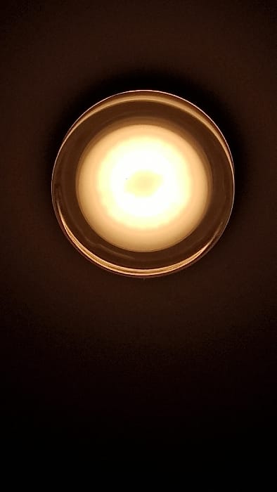 The image displays a single, lit tealight candle contained within a clear, cylindrical glass holder, viewed from a direct overhead perspective. The white candle wax emits a bright, warm yellowish-white light from its central wick, dominating the upper portion of the frame. The immediate environment consists of a uniform, dark background, appearing black or very dark brown, with no other discernible objects, individuals, or shadows visible. This scene is located in Florenville, Belgium.