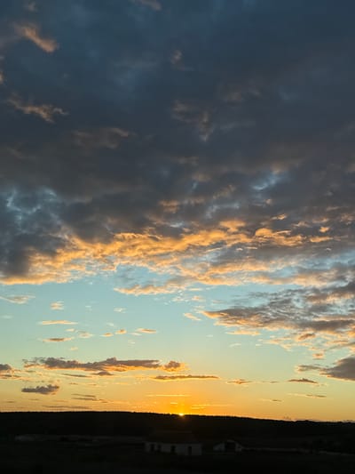 The image depicts a sunset over a rural landscape, possibly in the vicinity of Большое Тябердино, Russia. Dark, heavy clouds dominate the upper portion of the sky, tinged with orange and pink hues where they catch the setting sun's light. Below these, lighter, scattered clouds are illuminated by the sun, creating a gradient of color from a pale blue to a vibrant orange near the horizon. The sun itself is a bright orb just above the tree line. The foreground is a dark silhouette of a treeline and some indistinct structures, likely buildings, at the bottom. The overall impression is one of a dramatic and colorful sunset in a natural, undeveloped setting.