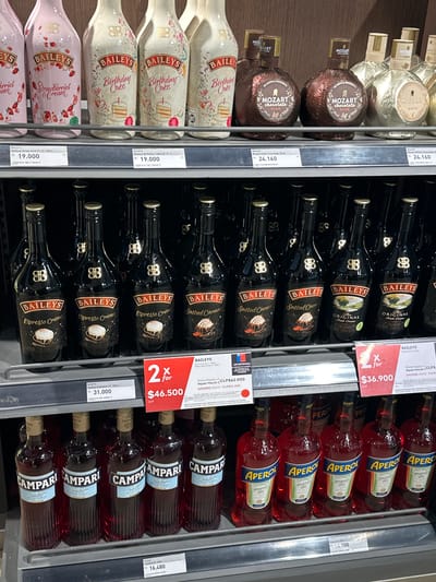 This is a photograph of a refrigerated shelf in a store. The products are all alcoholic beverages, specifically liqueurs and aperitifs.

From left to right, the products are:
*   Mozart Chocolate Liqueur (White and Dark)
*   Bailey's (Strawberry & Cream, Birthday Cake, Original Irish Cream, Salted Caramel, Espresso Crème)
*   Aperol Aperitivo
*   Campari

The prices for the products are visible on shelf tags. Some items have special pricing, such as "2 for" promotions. The currency is CLP (Chilean Peso).

The image was taken in Pudahuel, Chile.