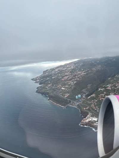 A sideways aerial view from an airplane window shows a coastal landscape in Santa Cruz, Portugal. The edge of the wing is visible in the upper right corner. The landmass is densely populated with buildings and vegetation. The coastline is rugged with cliffs meeting the ocean. A misty, overcast sky is to the left.