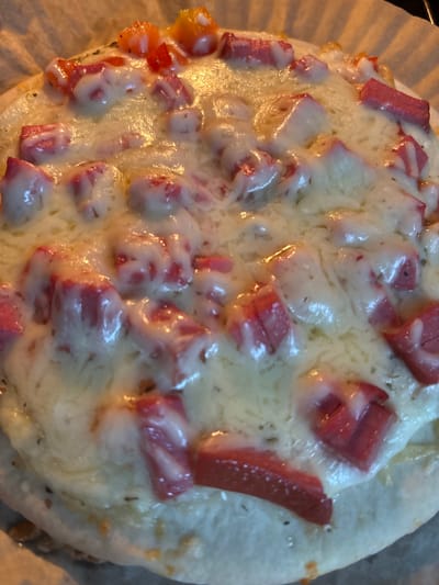 This is a close-up, overhead shot of a pizza baking on parchment paper. The pizza has a white crust and is covered in melted white cheese, diced pieces of red and pink meat, and some small red chunks that appear to be tomatoes. The lighting is warm and bright, creating highlights on the cheese. There is no indication of the city of Imeni Morozova, Russia in this image.