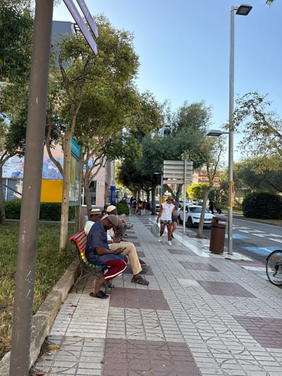 The image depicts a street scene in Tossa de Mar, Spain. In the foreground, three men are seated on a colorful park bench. The man on the left wears a blue patterned shirt and a grey cap, while the man in the middle is in a light-colored shirt and a white patterned hat. The man on the right is wearing a navy blue shirt with maroon and white striped pants, and black flip-flops. He is looking down at a mobile phone. The bench is positioned on a sidewalk paved with square tiles in various shades of grey and brown.
Behind the bench, a grassy area is visible, bordered by a low stone wall. Beyond the grass, a building with a mural painted on its exterior can be seen. Trees with green foliage line the street.
Further down the sidewalk, several people are walking. Two individuals are prominent: one wearing a white tank top and patterned shorts, carrying a large plastic bag, and another person walking alongside them. In the background, cars are parked and driving along the road, which has blue painted parking zones. A streetlamp stands tall on the right side of the sidewalk. A trash bin is also present on the sidewalk near the streetlamp. The sky is clear and blue.