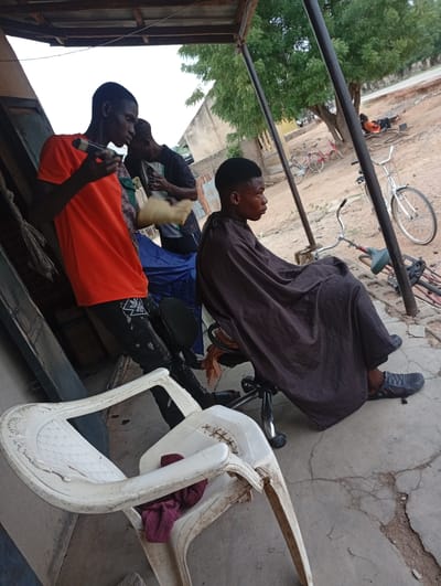 A young man, likely a barber, is holding clippers and preparing to cut the hair of another young man sitting in a barber's chair. Both are on a dusty, concrete ground, likely in a makeshift outdoor barber shop in Enagu, Nigeria. A few bicycles are parked in the background, and a large tree provides shade. In the foreground, two white plastic chairs are visible. The overall scene appears to be a casual, everyday moment in a Nigerian town.
