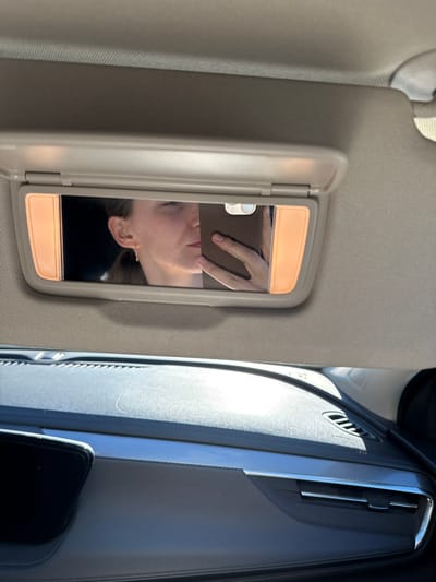 The image captures a close-up view of a car's interior, focusing on the passenger-side sun visor and a portion of the dashboard. The sun visor is open, revealing a vanity mirror with integrated lights. Reflected in the mirror is a person's face, oriented upside down, and a hand holding a smartphone. The person appears to be looking at the phone. The lighting in the mirror is warm, likely from the vanity lights.

To the right of the sun visor, the car's dashboard is visible, featuring a dark, textured surface and an air vent with chrome detailing. The sunlight is casting a strong glare on the dashboard, suggesting it is daytime. The overall scene is consistent with being inside a car, possibly during a sunny day. No visible text is present in the image. The location context provided is Barysaw, Belarus.