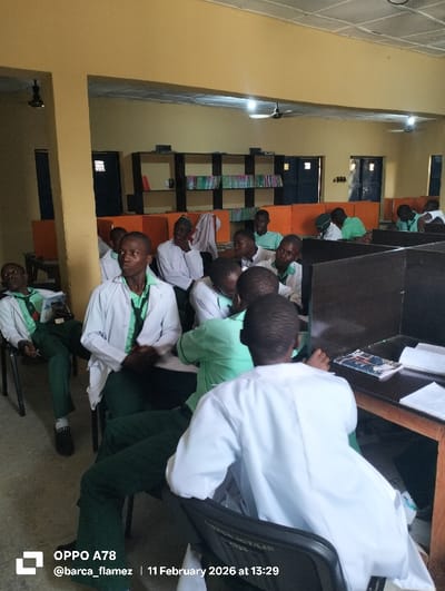 The image, captured on February 11, 2026, at 13:29, depicts an indoor library or study hall in Rinze, Nigeria. Approximately 15 to 20 young males, presumed to be students, are seated on black chairs at wooden tables. The attire primarily consists of white lab coats worn over light green shirts and dark green trousers, though some individuals wear only the green shirt and trousers.
The room features light yellow walls and a lighter ceiling equipped with overhead fluorescent lights and dark ceiling fans. In the background, dark-framed bookshelves stocked with numerous books line the far wall. Dark doors and windows with horizontal bars are also visible. Several tables incorporate orange privacy partitions, creating individual study carrels.
Students are observed in various postures: some face generally forward, some are turned to the side, one student in the far left of the frame is slumped in his chair with his eyes closed, and another in the immediate foreground is seated with his back to the camera. Books and papers are discernible on some of the desks.