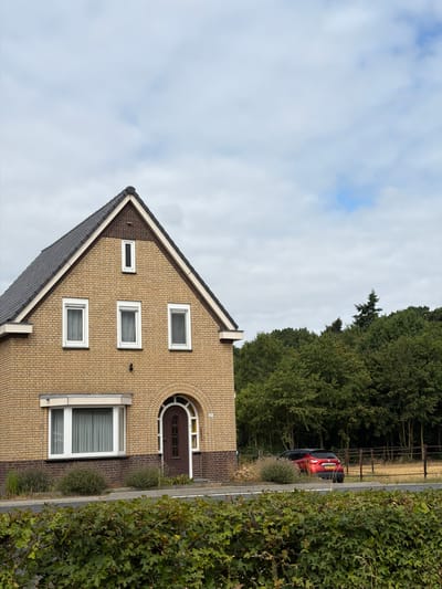 The image shows a two-story brick house with a dark grey tiled roof. The house is situated in the city of Melick, Netherlands. The front of the house features several white-framed windows of varying sizes, with the largest window on the ground floor being a bay window. A dark brown arched door is visible on the ground floor, to the right of the bay window. The house is surrounded by greenery, with a dense hedge in the foreground and a treeline in the background. A red SUV with a Dutch license plate is parked on the right side of the image, beyond a fence. The sky is overcast with scattered clouds.