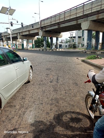The image shows a street intersection in Ado Ekiti, Nigeria, at coordinates 7.63 latitude and 5.214 longitude. A silver sedan is parked on the right side of the road, facing the camera. A person is sitting on a motorcycle, also on the right side of the road, looking to the left. The person is wearing a white shirt and blue jeans. The motorcycle is facing the camera. The intersection is under a concrete overpass with several columns. The traffic light in the middle of the overpass is red. There are buildings and trees in the background. The road is paved with asphalt. The day is clear and sunny.