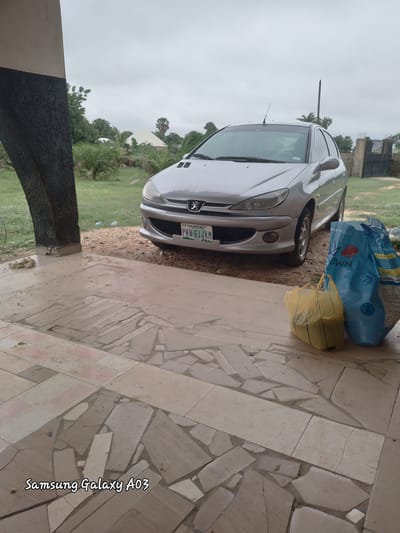 In Doka-Tofa, Nigeria, a silver Peugeot 206 sedan, license plate PKN-614KM, is parked on a dirt and gravel surface near a tiled patio. Two large bags, one blue and one yellow, are placed on the patio in front of the car. The scene is partially shaded by an overhead structure with a dark, textured pillar. In the background, lush green vegetation and trees surround some buildings, under an overcast sky. The tiled patio features a mosaic of irregularly shaped beige and brown tiles. The timestamp "Samsung Galaxy A03" is visible in the bottom left corner of the image.