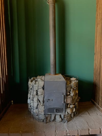 A cylindrical metal-caged sauna heater filled with irregular stones is centrally positioned on a raised brick platform in an interior space in Yasinia, Ukraine. The heater features a dark gray, rectangular metal firebox with a vertical door and a lower ash clean-out slot. A dark metal stovepipe extends vertically from the top of the firebox, showing signs of light rust at its base connection. Behind the heater, a solid dark green wall spans the background. To the left, a section of vertical wooden slats is visible, and to the right, a wider, stained wooden panel borders the scene. The brick platform beneath the heater extends forward, composed of reddish-brown pavers. No individuals are present.