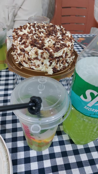 This media file displays a close-up, slightly angled view of a celebratory spread on a black and white checkered tablecloth. The primary subjects are a small, round cake heavily decorated with white and dark chocolate shavings, and a clear plastic cup containing a beverage with visible fruit slices and mint leaves. A large, green plastic bottle, possibly a soda, is partially visible in the background. A black plastic straw is inserted into the cup, and the lid of the cup has some embossed text and symbols. The cake is presented in a gold-colored plastic container. The overall scene suggests a casual gathering or personal treat. The lighting is soft, suggesting it could be daytime indoors or on a shaded patio. There are no people visible. The location context provided indicates Kara-Balta, Kyrgyzstan, but no specific visual cues within the image confirm this. Some embossed text is visible on the cup lid, but it is largely illegible. The green bottle has a partial label with some Cyrillic lettering, including "S" and "МОХИ".