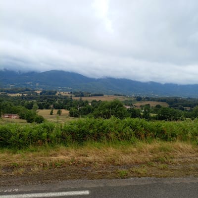 The image shows a cloudy, overcast sky over a mountainous landscape. In the foreground, there is a paved road with a white line marking. Beyond the road, there is a dense area of green ferns and dry grass. Further into the scene, rolling hills are covered with trees, with some fields and buildings scattered among them. In the distance, mountains are partially obscured by clouds. The architecture of the buildings suggests a rural or semi-rural setting. The image appears to be taken from a roadside perspective. There is no discernible specific location within the city of Cos, France, or any people present in the image.