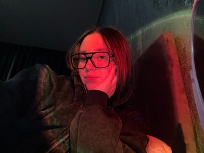 A single individual is depicted in a close-up, chest-up portrait. The subject, appearing female, has shoulder-length brown hair and wears black-framed glasses. Their left hand rests against the left side of their face, supporting the chin or cheek area. They are dressed in a dark, possibly charcoal grey, zippered hooded garment with a textured or faded appearance. The scene is dimly lit with atmospheric colored lights. Strong red light illuminates the subject's right side of the face (viewer's left), hair, and the adjacent background. A subtle greenish hue is visible on the left side of the subject's garment (viewer's right). The background consists of an interior setting: to the subject's right (viewer's left), dark fabric or drapes are visible, while to the subject's left (viewer's right), a dark, possibly upholstered or padded surface is present, with a textured wall behind it. The environment suggests an indoor space with mood lighting, possibly a lounge or similar venue.