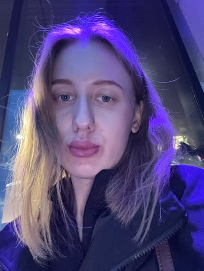 A selfie of a fair-skinned woman with blonde hair illuminated by purple lighting. The woman has blue eyes and a nose ring. She is wearing a dark jacket, possibly outdoors, in what appears to be an urban setting at night. The environment is dark with illuminated elements, possibly streetlights or other light sources reflected in a window or surface. There is no information about the location being Ol'ginskaya, Russia.