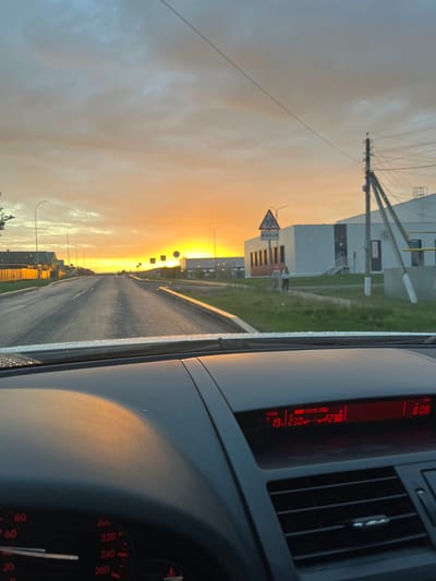 This is a first-person perspective from inside a vehicle driving on a paved road during a sunset. The sky is filled with dramatic orange and yellow clouds, casting a warm glow over the scene. The road ahead stretches into the distance, flanked by streetlights and buildings on either side. On the right side of the road, there is a modern white building with an entrance and stairs, and a utility pole with power lines. Two people are walking on a sidewalk in front of the building. A road sign with a triangular warning symbol is visible near the building. The car's dashboard is visible in the foreground, showing a digital display with temperature readings of 19 degrees Celsius outside and 23 degrees Celsius inside, along with the time 8:08. The car's speedometer is also partially visible, indicating a speed around 80. The overall impression is a drive through a suburban or rural area as day transitions into night.