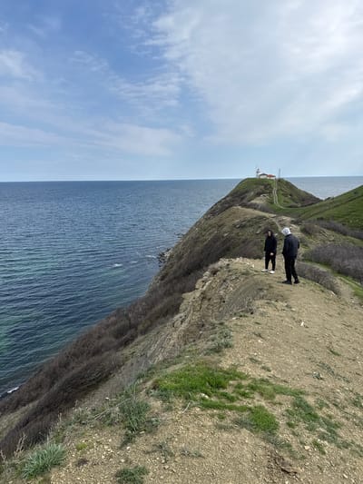 The image captures a coastal scene in Emona, Bulgaria, presenting a high-angle view of a cliffside overlooking the sea. Two individuals stand near the edge of the cliff, one wearing a white hoodie and dark clothing, and the other in dark attire. The cliff face exhibits a blend of exposed earth, rocks, and sparse green vegetation. In the distance, a building resembling a lighthouse or station is visible atop a grassy hill, connected by a path. The sky is partly cloudy, and the sea stretches to the horizon.