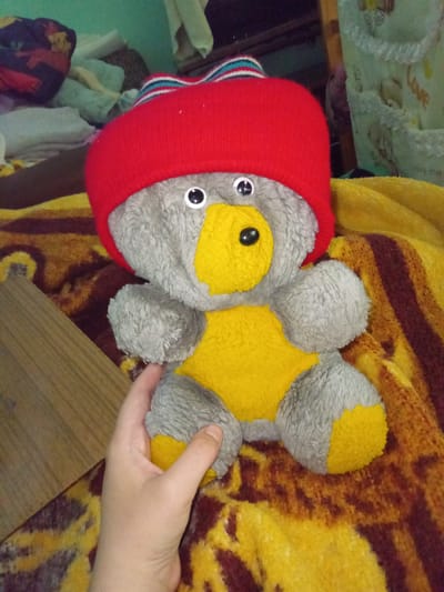 A stuffed teddy bear with gray fur and a yellow belly and feet wears a red knitted hat with a striped brim. The bear is sitting on a brown and yellow striped blanket. A human hand is visible holding the bear's left arm. In the background, there are various soft objects, possibly bedding or clothing, and a wooden surface. The scene appears to be indoors, in a domestic setting.