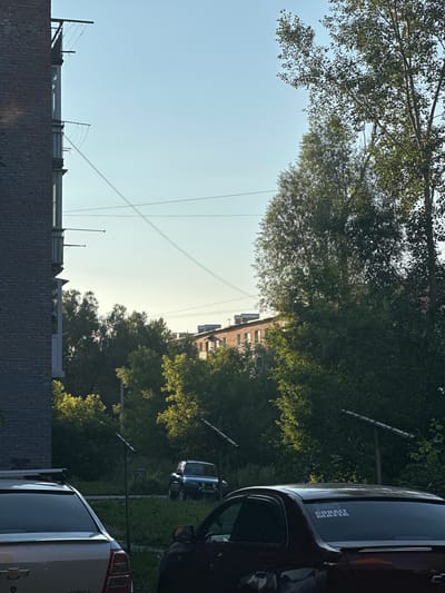 This is a vertical, slightly overexposed photograph of an outdoor scene, likely during the daytime, given the bright sky. The image is dominated by lush green trees on the right and top, with their leaves catching the sunlight. In the foreground, a dark brick wall runs horizontally across the bottom of the frame, obscuring the lower portion of the scene. Above the wall, a lighter-colored building with a modern, boxy design is visible, featuring metal supports and what appear to be ventilation units or decorative elements.
Several power lines crisscross the sky, suggesting an urban or semi-urban setting. In the middle ground, a row of buildings with a reddish-brown brick facade and white-trimmed windows can be seen nestled amongst the trees. To the right of these buildings, the rear of a dark-colored SUV is visible, with a sticker on the rear window that reads "COBALT MAFIA" with a stylized car graphic. Further to the right, the back of a silver vehicle, possibly another SUV or van, is also partially visible.
The overall impression is a snapshot of a residential or mixed-use area with significant greenery. The light suggests it's a sunny day, possibly late afternoon or early morning, given the angle of the sun and the warmth of the light hitting the trees. There are no people clearly visible in the frame. The presence of the vehicles and buildings indicates a developed area. The location context provided is Ridder, Kazakhstan.