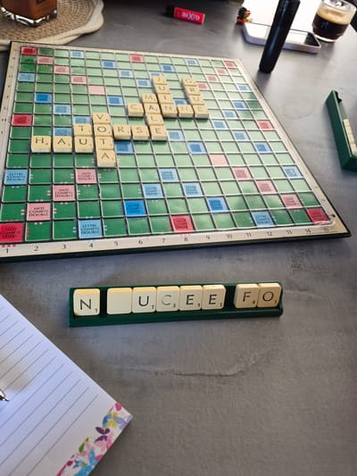 A Scrabble game in progress sits on a grey surface, with the board displaying a partially completed word arrangement. To the right of the board, a letter rack holds the tiles N, U, C, E, E, F, and O. A lined notebook with a floral design, topped with a pen, is positioned in the lower right corner. In the background, there's a dark drink in a glass, a white electronic device, and a black object (possibly a pen or marker). The game is likely located indoors, but the specific location within Villerupt, France cannot be determined from the image. The focus is on the game itself and the associated stationery, with other items scattered in the periphery.