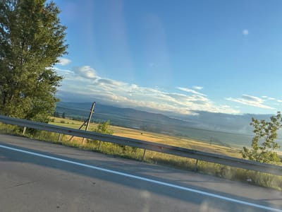The media file depicts a scenic view from a moving vehicle, likely on a highway, overlooking a rural landscape. The perspective is tilted, suggesting the photo was taken from a car window. The foreground features the asphalt road with a white dividing line, a metal guardrail, and foliage from trees. Beyond the guardrail, a vast field stretches out, illuminated by the golden light of what appears to be late afternoon or early evening. In the distance, rolling hills and mountains are visible under a bright blue sky dotted with scattered white clouds. A utility pole with wires can be seen in the middle ground. The overall impression is one of a peaceful journey through the countryside. The location is identified as Kvemo Rene, Georgia, hinting at a European setting. No people or specific activities are evident, but the scene evokes a sense of tranquility and natural beauty. The time of day is suggested by the warm lighting, and the weather appears clear and pleasant.