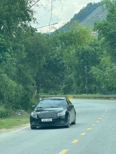 A black sedan, license plate number 30V-4698, is parked on the side of a road marked with dashed yellow lines. The surrounding environment features dense green vegetation, and a distant mountain slope is visible. The scene is located in Hà Giang 2 Ward, Vietnam.