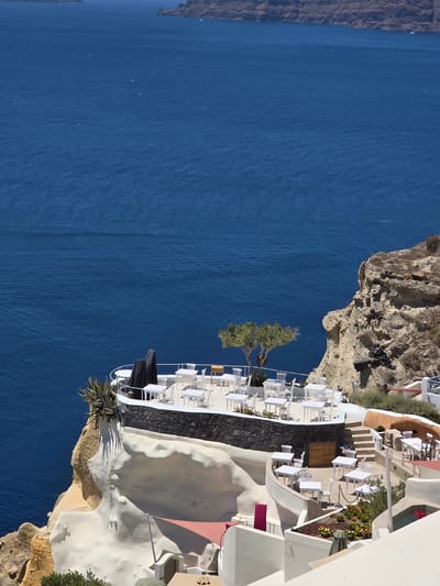 This media file depicts a scenic view of a white, multi-tiered restaurant or cafe perched on a cliff overlooking a vast expanse of deep blue water. The architecture is characteristic of Mediterranean island style, with whitewashed walls, curved structures, and numerous outdoor seating areas furnished with white tables and chairs. A single, sparsely-leaved tree stands prominently near the edge of the cliff, its branches reaching out over the water. Lush green vegetation, including an agave plant, grows around the structure and on the rocky cliff face. In the distance, the rocky terrain of the coastline is visible. The water displays subtle wave patterns, suggesting a calm day. The lighting indicates it is daytime, likely with clear skies. The overall impression is one of serene beauty and a tranquil setting ideal for dining or relaxation, with a clear view of the sea. There is no visible text in the image.