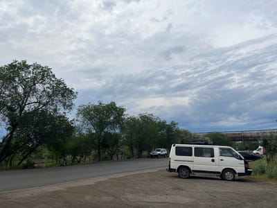 A white van is parked on a dirt shoulder next to a paved road in an industrial area. In the background, a white SUV is parked on the side of the road, and further back, other vehicles and industrial infrastructure, including a large pipe suspended on supports, are visible. Lush green trees line the opposite side of the road. The sky is overcast with textured clouds. The scene appears to be in the city of Temirtau, Kazakhstan.