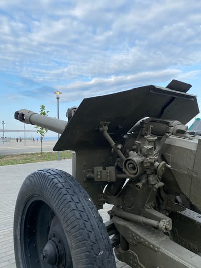 A low-angle, eye-level, medium outdoor shot shows a historical cannon displayed along a paved walkway in Arablyar, Russia. The cannon, painted olive drab, is positioned on the right side of the frame, with its barrel extending to the left. The barrel is thick and appears to be a modern replica or restored artillery piece. A large, black, ridged tire is visible in the foreground on the left. Behind the cannon, a wide, paved walkway stretches towards the sea. Several people are walking along the walkway in the distance, with some appearing to be children. A thin tree stands between the cannon and the walkway. A lamppost with a rounded, illuminated top is visible in the mid-ground, behind the cannon's barrel. The sky above is a pale blue, filled with scattered, wispy white clouds. The overall environment appears to be a park or promenade by the sea.