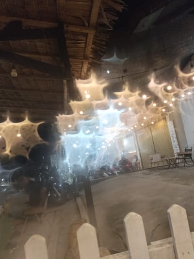 This media file depicts an outdoor scene at what appears to be a cafe or open-air establishment, likely in Blangbitra, Indonesia. The setting features a thatched roof structure supported by wooden beams, suggesting a rustic or tropical aesthetic. String lights are strung across the area, many of which are illuminated, casting a warm glow. Some of these lights are surrounded by a hazy, starburst effect, possibly due to motion blur or atmospheric conditions.
In the foreground, a white picket fence is partially visible, and beyond it, the ground is paved or a packed dirt surface. In the mid-ground, there are several motorcycles parked, indicating a place where patrons might arrive by vehicle. Some tables and chairs are set up, suggesting dining or lounging areas. A building with large doors, possibly a shop or part of the establishment, is visible in the background.
The time of day appears to be evening or night, given the artificial lighting and the dark sky. The overall mood is relaxed and inviting, with the soft lighting creating a cozy ambiance. There is a slight blur and haziness throughout the image, which could be due to low light conditions, motion, or the camera's focus.
The image captures a moment of stillness in what seems like a communal gathering space. There is a suggestion of people present, with dark figures visible among the motorcycles, though their details are indistinct due to the lighting and blur. One figure in the left mid-ground is particularly noticeable, leaning forward.
On a white board or sign on the right side of the background building, there is some text that appears to read "DELLON," suggesting the name of the establishment or a brand. The weather is not explicitly indicated, but the outdoor setting implies it is suitable for being outside.