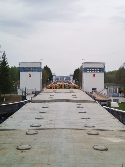 The image shows a canal lock, likely the 5th lock in a series. Two symmetrical, white buildings marked with a large red "5" stand on either side of the canal. A concrete bridge or platform with several round hatches stretches across the foreground, providing a high-angle view into the lock system. Various pieces of machinery and equipment are visible between the buildings. Trees and greenery surround the area, suggesting a location in a natural or park-like setting. The sky is overcast. The city of Alekseevskoe is not discernible from the image alone.