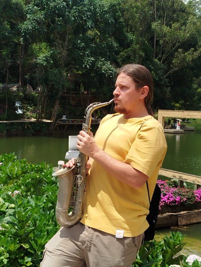 A Caucasian man with long brown hair and a goatee plays a saxophone outdoors. He is wearing a yellow t-shirt and tan cargo shorts. The man is positioned in the foreground, with a body of water and lush greenery behind him. In the background, there are trees, a wooden boardwalk with people, and a small building. The overall environment suggests a park or natural area. The image appears to be taken during the day, with bright lighting.