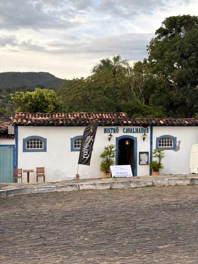The image captures the "Bistrô Cavalhadas" restaurant in Pirenópolis, Brazil. The building is white with blue trim around the windows and doorway. A black flag with white lettering hangs near the building's roof. The restaurant's name is written in blue above the entrance. To the side of the entrance, a sign advertises "Almoço Executivo" (Executive Lunch). There are plants in pots near the entrance and by the windows. Wooden chairs and a table are situated outside the restaurant. The ground is covered in cobblestones. Green trees and a hill are visible in the background, with a partly cloudy sky.
