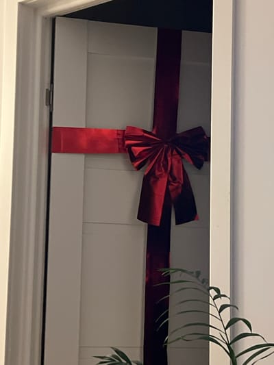 An indoor shot shows a closed white door decorated with a large red ribbon tied in a bow. The ribbon is positioned vertically and horizontally across the door, resembling a gift wrap. The door appears to be made of wood and has horizontal paneling. To the right of the door, a portion of a white wall is visible. In the lower right corner, a few green leaves of a plant are peeking into the frame. The scene is dimly lit, suggesting it might be evening or a poorly lit room. No individuals are present in the image, and no specific details about Florești, Romania, are discernible from the visual information provided.