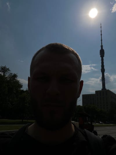 The image is a selfie taken outdoors, with the sun directly overhead and slightly to the right, causing the foreground subject's face to be heavily silhouetted and largely obscured. The subject, a person with short hair and a beard, is positioned in the center of the frame, facing forward. Behind the subject, the sky is blue with scattered white clouds. A tall, slender television tower with a pointed top is visible in the upper right quadrant of the image, with a building of similar height situated to its left. The lower portion of the image shows a street with a sidewalk, trees, and some parked vehicles. The general setting appears to be an urban or suburban environment in Вёшки, Russia.