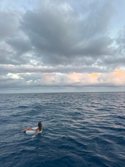 The image displays an individual floating in the open Pacific Ocean, off the coast of East Honolulu, United States. A single adult female, with long dark hair, is visible from behind and to her left, supported by a white, circular inflatable ring. She is partially submerged in the water from the chest down, wearing a light-colored swimsuit, and oriented towards the horizon. The water is a deep blue with visible surface waves and ripples. The sky above is largely covered by dense, grey cloud formations, with subtle faint pink and orange hues present on the undersides of some clouds along the distant horizon line, particularly towards the right side of the frame. No landmasses or other subjects are visible.