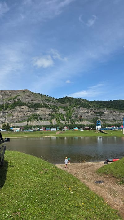 The image depicts a scenic riverside view in the village of Tash-Asty, Russia. A calm body of water, likely a river, dominates the foreground, reflecting the blue sky and clouds. On the far bank, a small village is nestled against a large, layered rock formation and lush green hills. The village consists of several colorful houses with red, blue, and green roofs. A white suspension bridge with blue supports spans across the river near the village. In the foreground, a grassy bank slopes down to the water. Two children, one wearing a hat and the other in a green shirt, are near the water's edge, with a raft lying on the bank to the right. A dark-colored vehicle is partially visible on the left side of the frame. The scene suggests a peaceful, rural setting on a bright, sunny day.