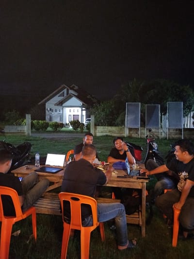 In Tanjongteungkuali, Indonesia, at night, a group of five men are gathered around a wooden table outdoors. They appear to be engaged in a discussion or collaborative work session, with several laptops open on the table. Two men are looking at their laptops, one is looking away from the table, another is looking at the table and gesturing with his hands, and the fifth man is looking at his laptop. Two of the men are wearing black t-shirts, and the other three are also dressed in casual dark clothing.

The environment is dimly lit, with the light source appearing to be the laptops and possibly some overhead lighting not visible in the frame. A white house with a distinctive roof structure is visible in the background, along with some neatly trimmed bushes and trees. The ground is covered with grass. Several motorcycles are parked nearby, one of which is partially visible in the foreground. The overall atmosphere suggests a relaxed yet focused gathering.
