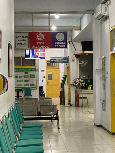 This is an indoor, eye-level, portrait-oriented photograph of a hospital waiting area in Margoyoso, Indonesia. The scene is illuminated by artificial light, suggesting it is likely evening or indoors without natural light.
The primary subjects are the objects and the setting itself. The waiting area appears clean and functional, with tiled walls and floors. A row of green padded benches for seating is prominently displayed in the foreground, some of which are empty. Behind the benches, there are stacks of what appear to be metal waiting chairs.
Several signs are visible, providing information and instructions. A dark blue sign with a smiley face reads "UDARA SEGAR" (Fresh Air). Below it, a red sign with a cigarette crossed out states "STOP MEROKOK" (Stop Smoking). Further down, a sign reads "KAWASAN TANPA ROKOK" (No Smoking Area). Another sign in white with green text, oriented towards the back of the room, says "PASIEN RAWAT JALAN RSI PATI" (Outpatient Department RSI PATI). A smaller blue sign with white text above a yellow door says "PELAYANAN INFORMASI" (Information Service).
In the background, a yellow double door is visible, along with a fan and a bouquet of flowers on a ledge. There is also a television screen showing an image of what appears to be a man in a suit, possibly a news broadcast or informational video. Further into the hallway, a number "3" is visible on a sign. A wheelchair is parked near the yellow door. The overall impression is a functional and orderly public space within a healthcare facility.