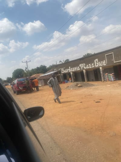 The scene is set outdoors in Tataura, Nigeria, under a bright, partly cloudy sky. The perspective is from within a dark-colored vehicle, with its side mirror visible in the lower left foreground.
An unpaved, reddish-brown dirt road occupies the mid-ground. On the left side of this road, a red SUV or truck is parked, its rear tailgate open or damaged, exposing internal components. Three individuals are gathered around the vehicle's rear; one wears an orange shirt, and another a dark shirt.
In the center-right of the mid-ground, a man in a light grey traditional long tunic and dark shoes walks from left to right across the dirt road, carrying a dark briefcase in his left hand.
On the right side of the road, the "Sardauna Plaza Gwt" building is visible. It is a single-story, light grey structure featuring multiple commercial bays. A pile of dirt and rocks is located in front of the plaza, to the right of the walking man. Stacks of bottled or canned beverages are arranged outside one of the plaza's bays on the far right.
In the background, a wooden utility pole with multiple overhead power lines is present on the left. Green trees and additional structures are visible behind the vehicles and buildings.