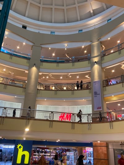 This media file captures an interior view of a multi-level shopping mall. The scene is brightly lit with artificial lights, suggesting it could be daytime or evening. The architecture features curved walls and multiple levels with railings, creating an open atrium effect.
Several people are visible on different levels, walking along the walkways and near shop entrances. Some are casually dressed, implying a typical shopping day. There are various signs and advertisements displayed throughout the mall, indicating retail establishments. Notable visible text includes "H&M," "GET HK GE&M HOUPON," "HELLO, HUT STUFF," "MORE D177A," and "JAYA." The presence of these brand names and messages reinforces the commercial nature of the setting.
The mall appears to be a busy commercial hub, with shoppers navigating the various floors. The overall atmosphere seems neutral, focusing on the architectural details and the retail environment. The specific location is identified as Pekanbaru, Indonesia, which provides context for the cultural and architectural style. The media file does not provide information about the weather or time of day, but the indoor setting makes these less relevant.