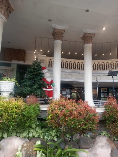 The scene depicts the outdoor entrance area of a building in Suddepalle, India, adorned with festive decorations. In the foreground, a landscaped garden features multiple large, smooth grey rocks interspersed with various shrubs. These include dense green foliage, smaller shrubs with distinctive reddish-green leaves, and light green ground cover plants.
Above this garden, a set of light grey steps leads to a raised patio area. On the left, a large white ornate planter contains green foliage. Central to the patio is a tall, artificial green Christmas tree positioned beside a large Santa Claus figure. The Santa Claus figure is depicted in traditional red attire with white trim, black boots, a white beard, and holds a small green object in its left hand.
The building's facade is characterized by two prominent, white, fluted columns with ornate, possibly Corinthian, capitals. These columns support an overhead structure. Below this structure and between the columns, a curved white balustrade with multiple evenly spaced vertical elements forms a balcony or decorative railing. Hanging beneath the overhead structure and across the width of the scene are strands of string lights with exposed bulbs.
Behind the columns, the ground floor of the building is visible, featuring an entrance with dark-framed windows or doors, revealing a dimly lit interior. To the right of the rightmost column, a black metal music stand is positioned. The overall impression is one of a decorated public or commercial space. No living individuals are visible.