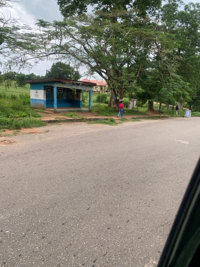 A medium shot captures a roadside bus stop on an overcast day in Ojere, Nigeria. The bus stop is a simple, open-air structure with a blue and white painted concrete building. It features a blue-tiled bench and an opening on the side facing the road. A white and blue emblem is visible on the left side of the bus stop, and a mural depicting Africa is painted on the right. The bus stop is surrounded by green grass and trees. In the foreground, a paved road stretches across the bottom of the frame. A person wearing a pink shirt and blue jeans walks away from the bus stop towards the right side of the frame. Further in the background, another person wearing a blue shirt and dark pants stands near a white sign. To the right of the bus stop, a building with a red roof is partially visible. The overall impression is of a quiet, rural bus stop.