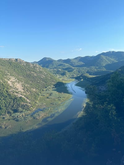 This is an eye-level, medium shot of a winding river flowing through a valley in Rvaši, Montenegro. The river is surrounded by lush green hills and mountains covered in trees. A small boat is visible on the river, leaving a white wake behind it. The sky is clear and blue, with the sun casting shadows across the landscape. The overall impression is one of natural beauty and tranquility.