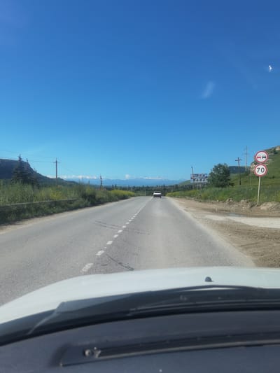 The image is taken from the perspective of a vehicle driver, looking forward on a paved road. The road has dashed white lines marking lanes and is bordered by grassy shoulders. In the distance, mountains are visible, some with snow-capped peaks. The sky is a clear, bright blue.

On the right side of the road, there are two circular traffic signs. The upper sign depicts two cars side-by-side, indicating a lane restriction. The lower sign displays the number "70" in a red circle, signifying a speed limit of 70. Behind the signs, there is a grassy slope with some scattered trees. A billboard is also visible in the middle distance on the right.

A white car is driving in the same direction on the road ahead. Vegetation, including tall green grass and some yellow wildflowers, lines the left side of the road. Power lines and poles are present along the side of the road.

The image appears to be taken in a rural or semi-rural mountainous region. While the prompt mentions Khabez, Russia, there are no specific landmarks or text in the image that directly confirm this location.
