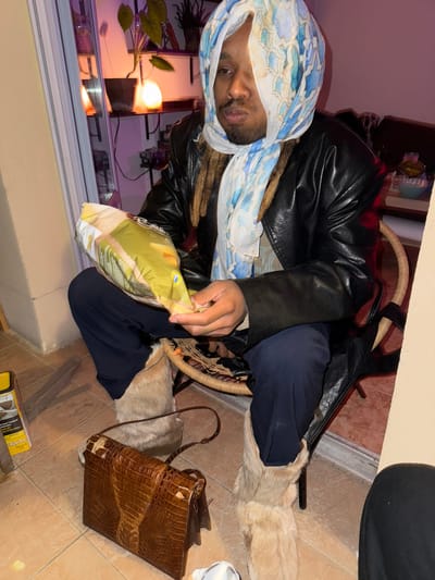A person wearing a blue and white head covering and a black leather jacket is seated in a wicker chair in an indoor environment. The person is holding a bag of chips. The person is wearing navy pants and light brown furry boots. Next to the person's chair is a brown alligator pattern purse. A box is on the floor nearby. The location could be Montreuil, France. There are plants and lighting visible near a window in the background.