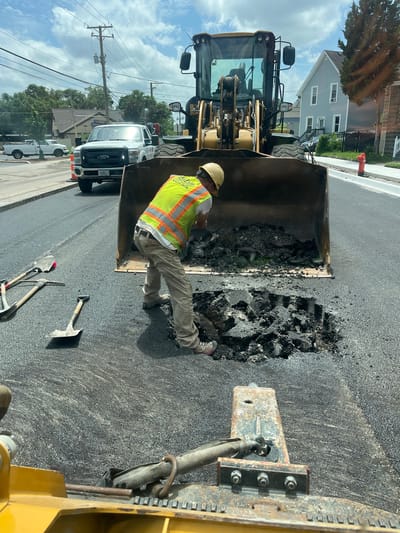 In Elgin, Illinois, USA, on a bright, partly cloudy day, a construction worker in a yellow high-visibility vest and hard hat is shoveling asphalt from the bucket of a yellow front-end loader into a large pothole on a paved street. Several shovels and a pickaxe lie on the asphalt near the pothole. A white Ford pickup truck is parked behind the loader, and houses are visible in the background, along with utility poles and wires. A red fire hydrant stands on the sidewalk. The scene appears to be a road repair operation.