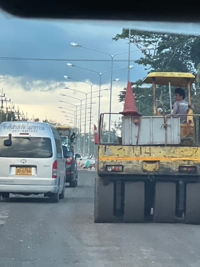 The image depicts a street scene in Tambon Don Sak, Thailand, characterized by a cloudy sky and a series of streetlights. In the foreground, a yellow road roller with a red cone on top is visible, with a person operating it. To the left of the road roller, a silver van with Thai license plates (30-0784) is parked, displaying Thai text on its rear door. Behind the van, a dark-colored car is present. The environment suggests ongoing road construction or maintenance.