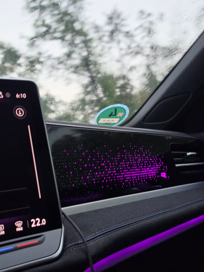 In Raunheim, Germany, the interior of a modern car is captured on a rainy day. The focus is on the dashboard, featuring two prominent digital displays. The display on the left shows the time "6:10" and a circular icon with a number "1" inside. Below this, part of a climate control interface is visible, displaying "22.0" degrees Celsius, with indicators for windshield defrost and maximum fan speed. To the right, a longer, wider display exhibits a dynamic pattern of purple illuminated dots, resembling a digital waveform or abstract graphic. Outside the car, the view through the rain-streaked windshield shows blurred trees and an overcast sky. A green circular sticker with some German text is affixed to the windshield, partially visible. Ambient purple lighting emanates from the lower dashboard area, complementing the illuminated display.