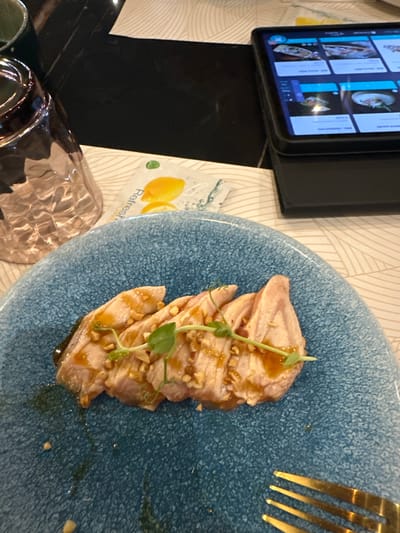 The image depicts a close-up view of a plate of sliced raw fish, possibly tuna or salmon, served on a textured blue plate. The fish is garnished with chopped nuts and a light green herb, and drizzled with a sauce. The plate is positioned on a table with a light-colored, patterned tablecloth. To the left, a partially visible glass and a dark object, possibly a phone or tablet, displaying images are present. In the background, a tablet screen is visible, showing a grid of food items. The scene appears to be indoors, likely a restaurant. The specific location in Ponteterra, Italy is not discernible from the visual information provided.