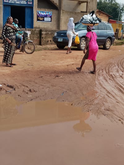 The image depicts a scene in Orudu-Olokan, Nigeria, characterized by a muddy, unpaved road with large puddles. In the foreground, a woman in a pink outfit and a leopard-print sash walks towards the right, carrying a basket of items on her head. Her reflection is visible in a large puddle. To her left, another woman in a black and white patterned dress stands near a motorcycle, with a man in a blue shirt riding it. Behind them, a blue SUV is parked. Further in the background, a building with blue doorways and a poster advertising "IGBIMO ASOMUBAYE DISTRICT" is visible. Another poster with pictures of individuals is also displayed on the building's exterior. The environment appears to be a semi-urban or rural setting, with a predominantly earthen landscape and vegetation in the distance.