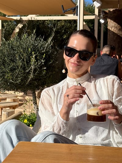 A woman is seated at an outdoor cafe table under a retractable beige canopy in the Anavissos Municipal Unit, Greece. She is positioned slightly to the left of the frame's center, facing forward with a slight smile. She wears black cat-eye sunglasses, a white high-collared blouse with a textured dotted pattern, and light-wash denim jeans. A long, thin gold-colored earring with a white pearl dangles from her right ear. Her left hand, adorned with a silver ring featuring a dark, possibly black, stone, holds a black straw inserted into a clear glass. Her right hand also grasps the clear glass, which contains a dark iced beverage with a layer of foam on top. The table in front of her is light brown wood. In the background, lush green olive trees are visible. To the far left, several stacked circular wooden stools are present, along with small yellow flowers. Behind the woman, a male figure is partially visible from the back, wearing a dark sleeveless top. Bright sunlight illuminates the scene, casting shadows.